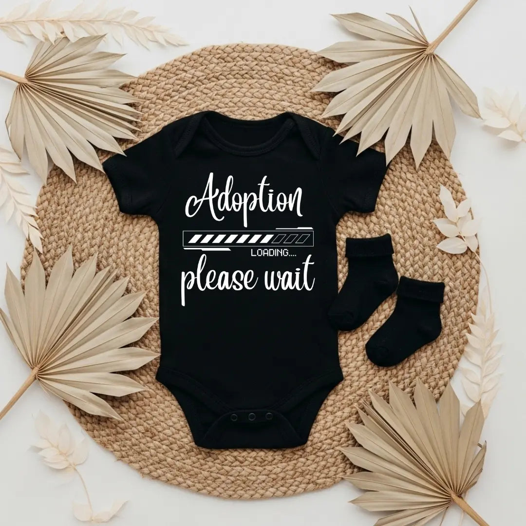White baby onesie with 'Adoption loading please wait' text on a textured surface with socks and dried leaves.
