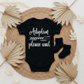 Black baby onesie with 'Adoption loading please wait' text on a woven mat with dried leaves.