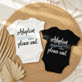 Two baby onesies with 'Adoption please wait' text on a woven mat.