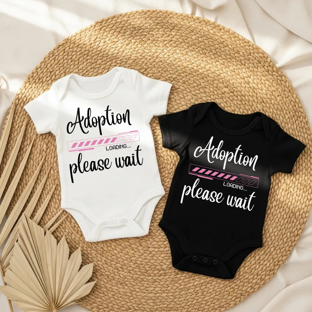 Two baby onesies with 'Adoption please wait' text on a woven mat.