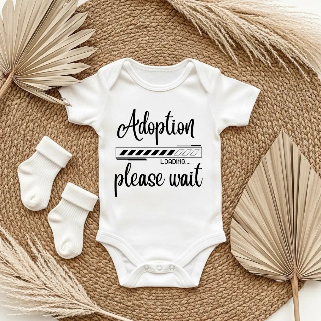 White baby onesie with 'Adoption loading please wait' text on a textured surface with socks and dried leaves.