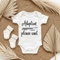 White baby onesie with 'Adoption loading please wait' text on a textured surface with socks and dried leaves.
