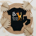 Black baby onesie with a deer design and text, placed on a woven mat with dried leaves.