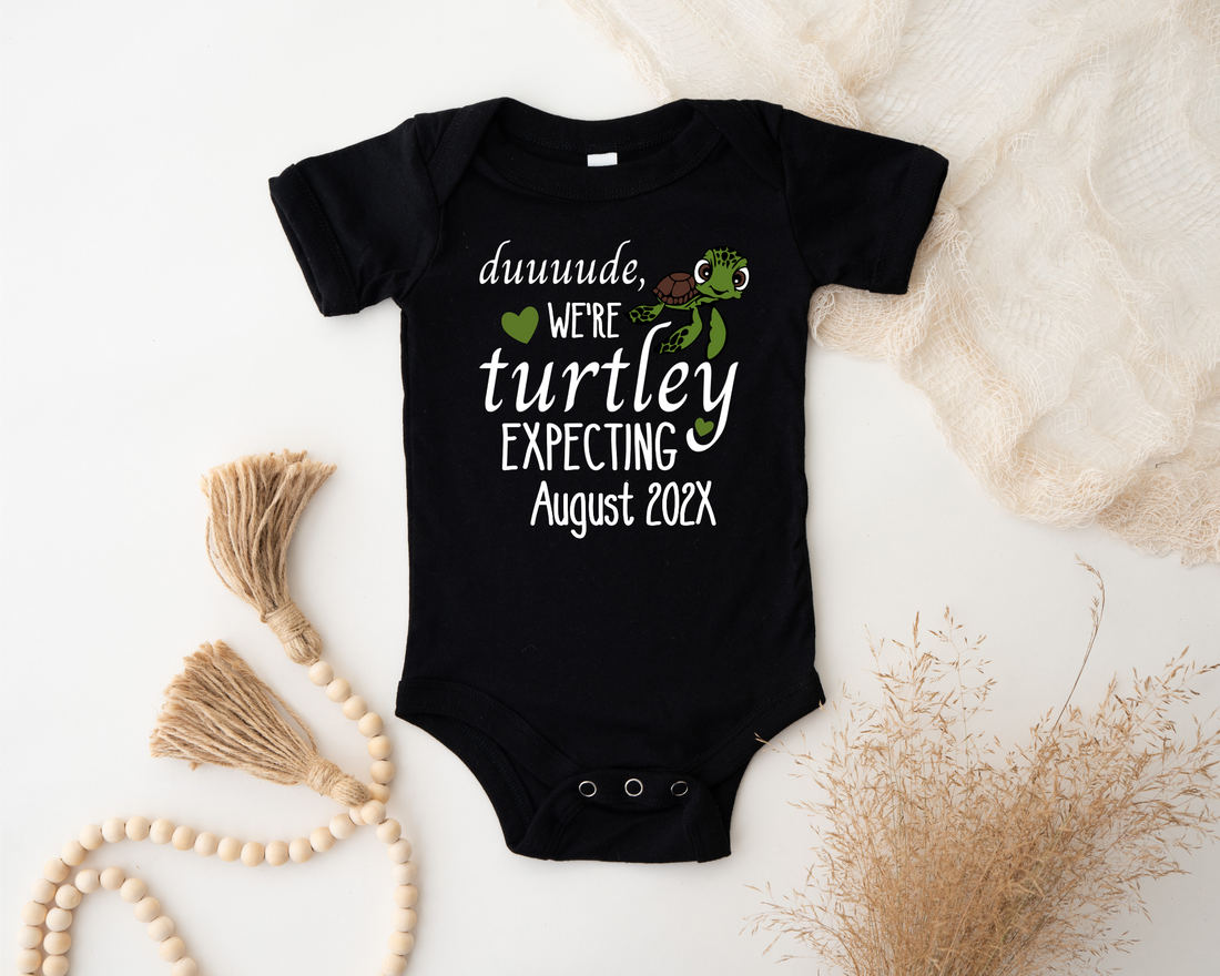 White baby onesie with turtle design and text on a textured beige surface.